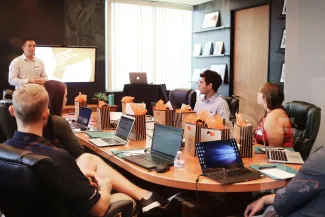 A group of diverse individuals seated around a table, each using a laptop, engaged in discussion or collaboration