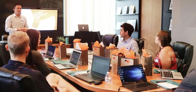 A group of diverse individuals seated around a table, each using a laptop, engaged in discussion or collaboration